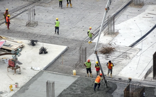 Robotnicy w kaskach i kamizelkach odblaskowych na placu budowy, wylewający beton na zbrojony fundament. Widok z góry.